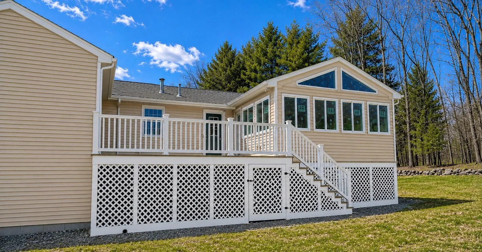 Tan vinyl-sided home with a raised rear deck, white railings and lattice skirting, and a sunroom bump-out with trapezoid transom windows