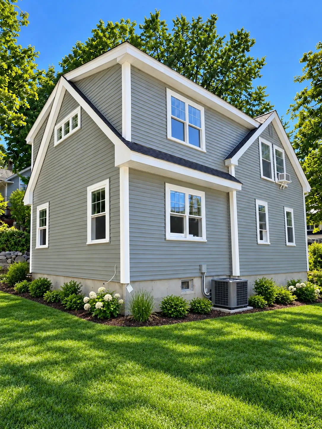Rear corner of a gray cape with a full-width shed dormer adding second-floor space, white trim, and foundation plantings