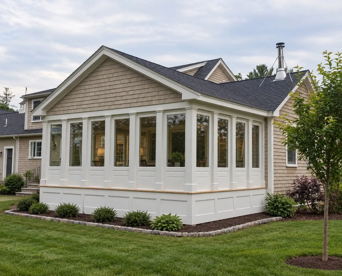 Sunroom addition with a cedar-shake gable, three walls of white-framed windows, a white paneled skirt, and a stone-edged planting bed
