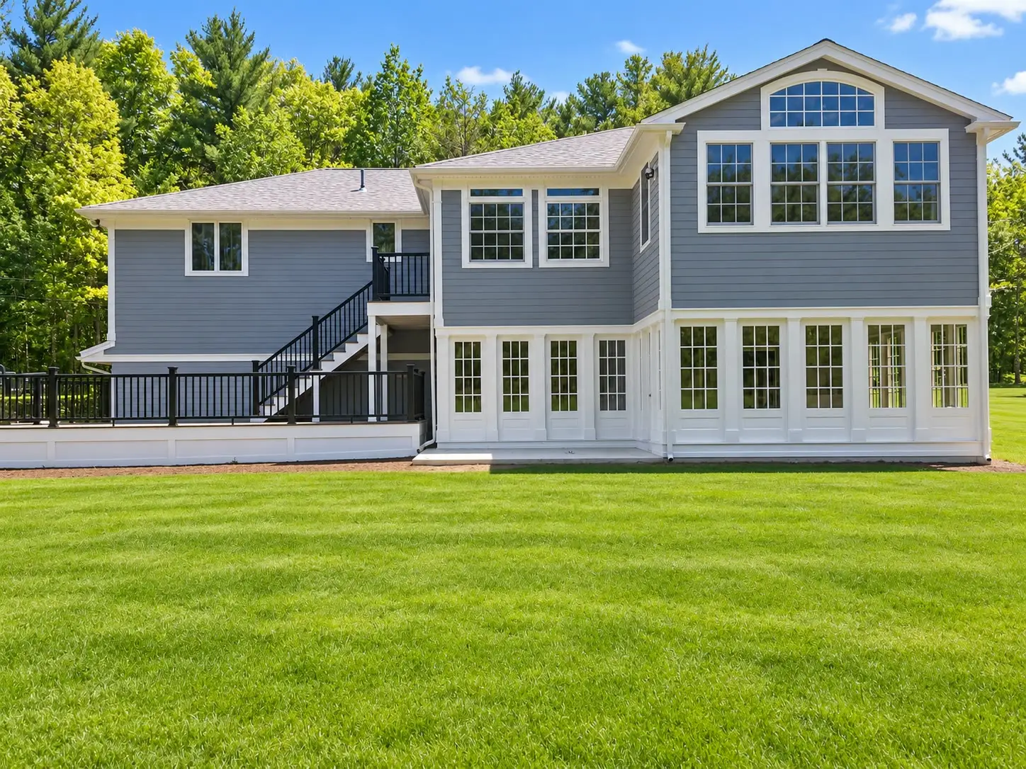Rear view of a gray two-story home with a white two-story addition featuring a wall of grid windows, an arched gable window, and a black-railed deck with stairs.