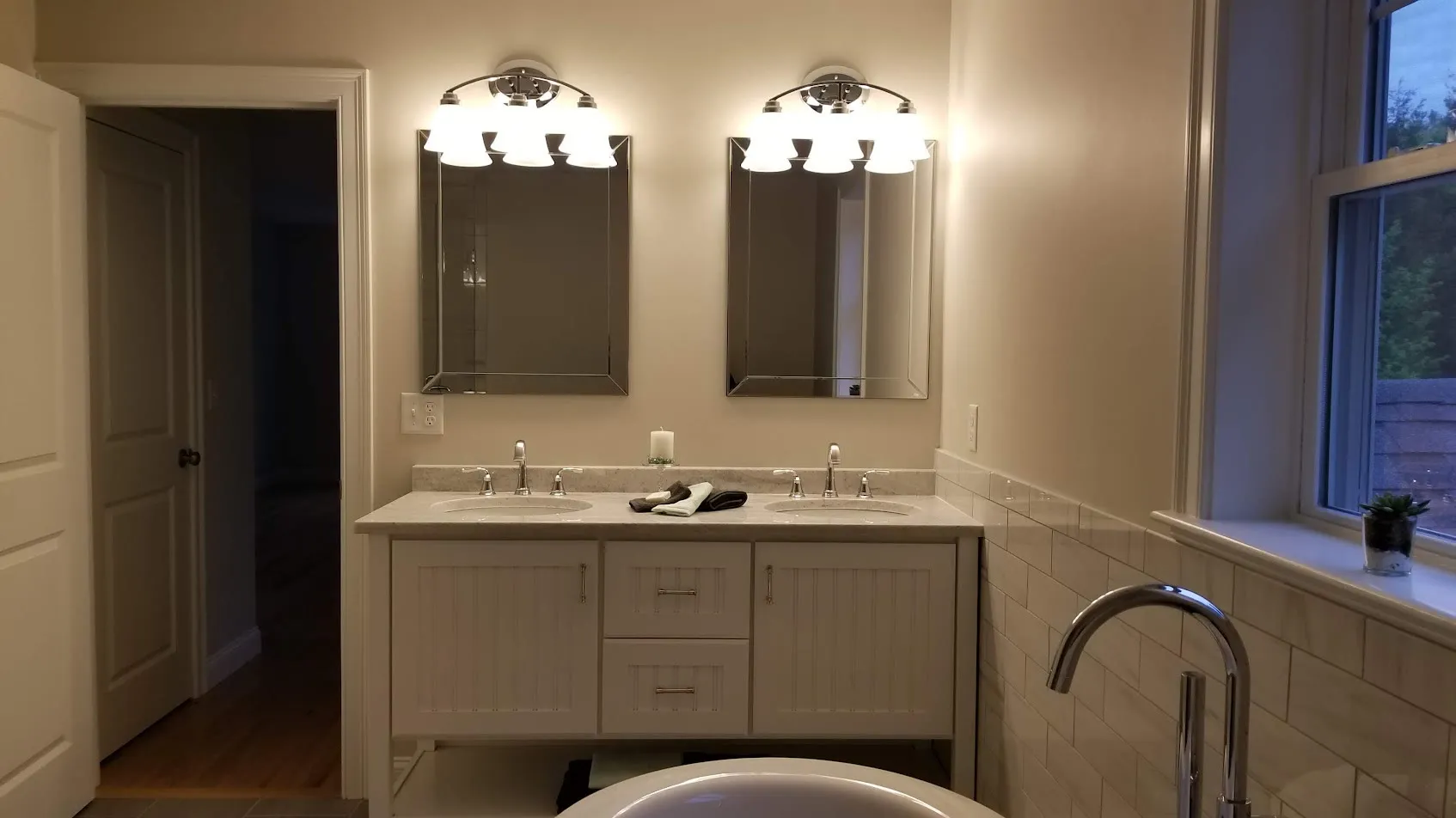 Warm-toned bathroom with a white beadboard double vanity, beveled mirrors, three-light fixtures, subway tile, and a soaking tub in a window alcove