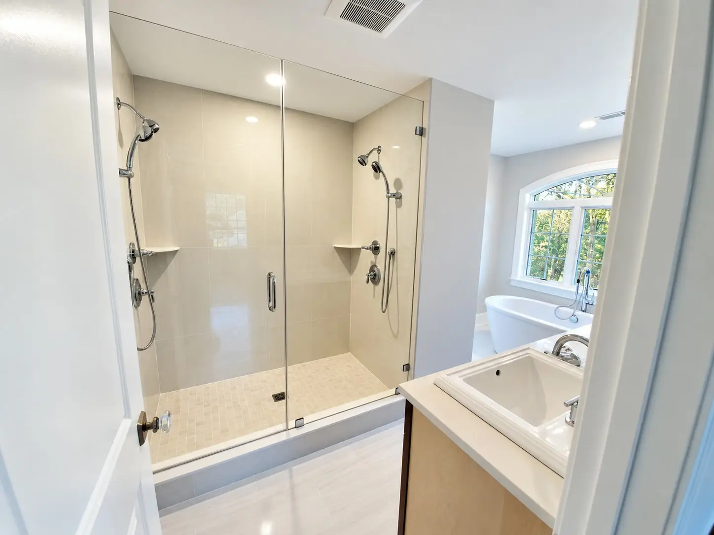 Glass double shower with dual showerheads on beige large-format tile, a maple vanity with a drop-in sink, an arched window, and a freestanding tub.