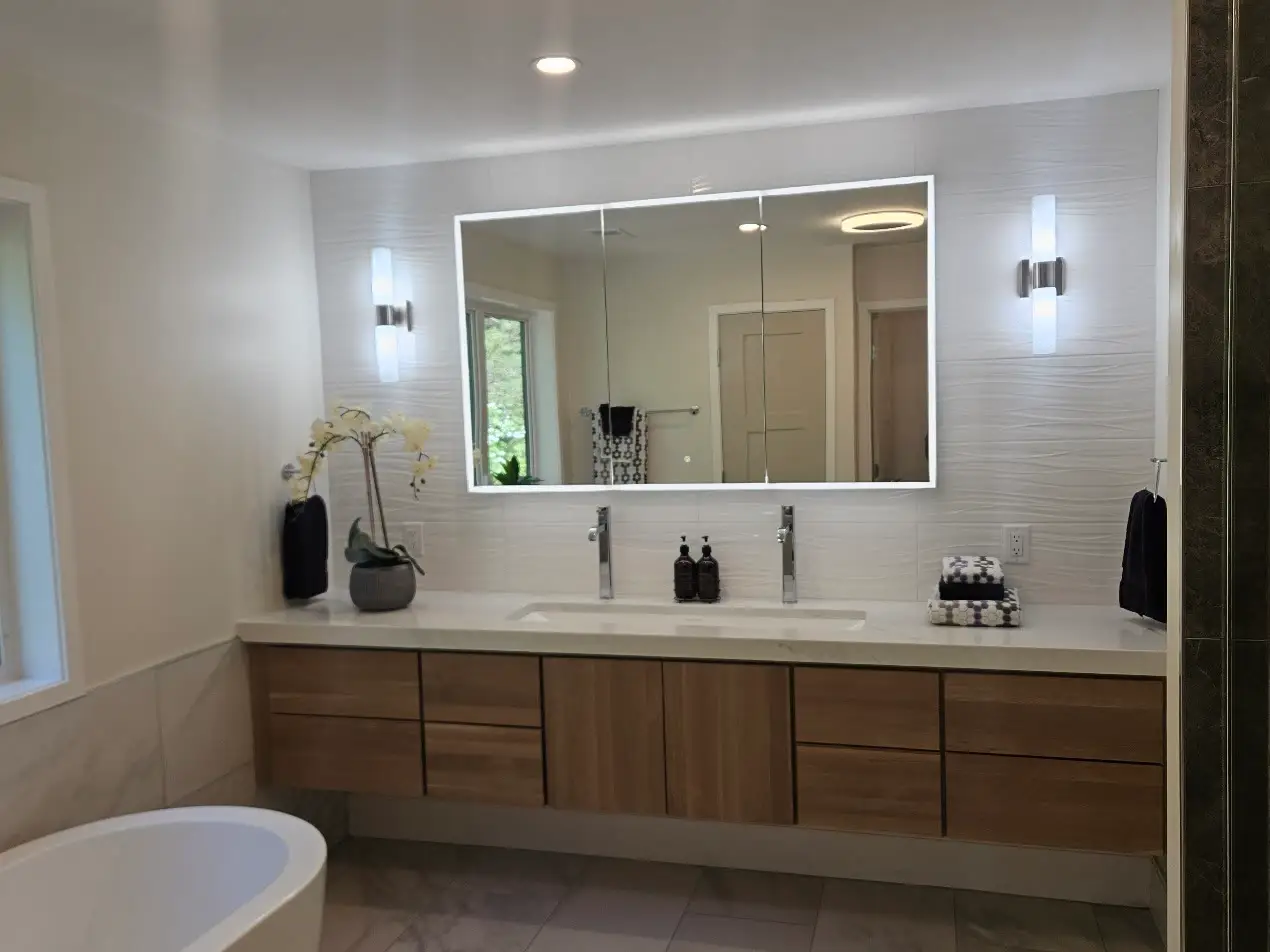 Floating walnut double vanity with a backlit mirror, white wave-textured tile, an orchid, and a freestanding tub in the foreground.