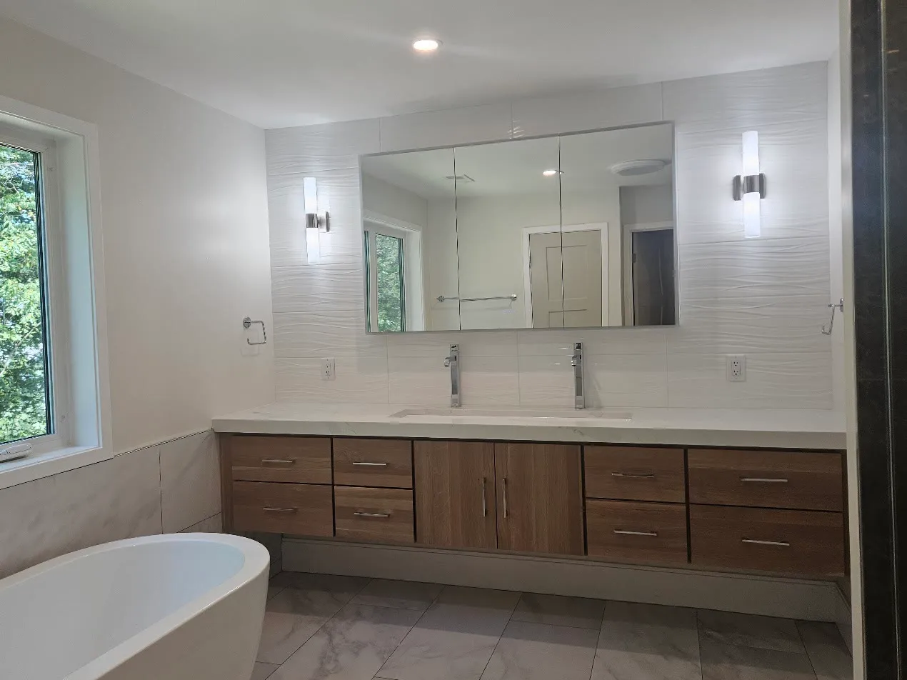 Modern bathroom with a floating walnut double vanity, wide mirror, white wave-textured wall tile, chrome sconces, marble-look floor, and a freestanding tub by the window