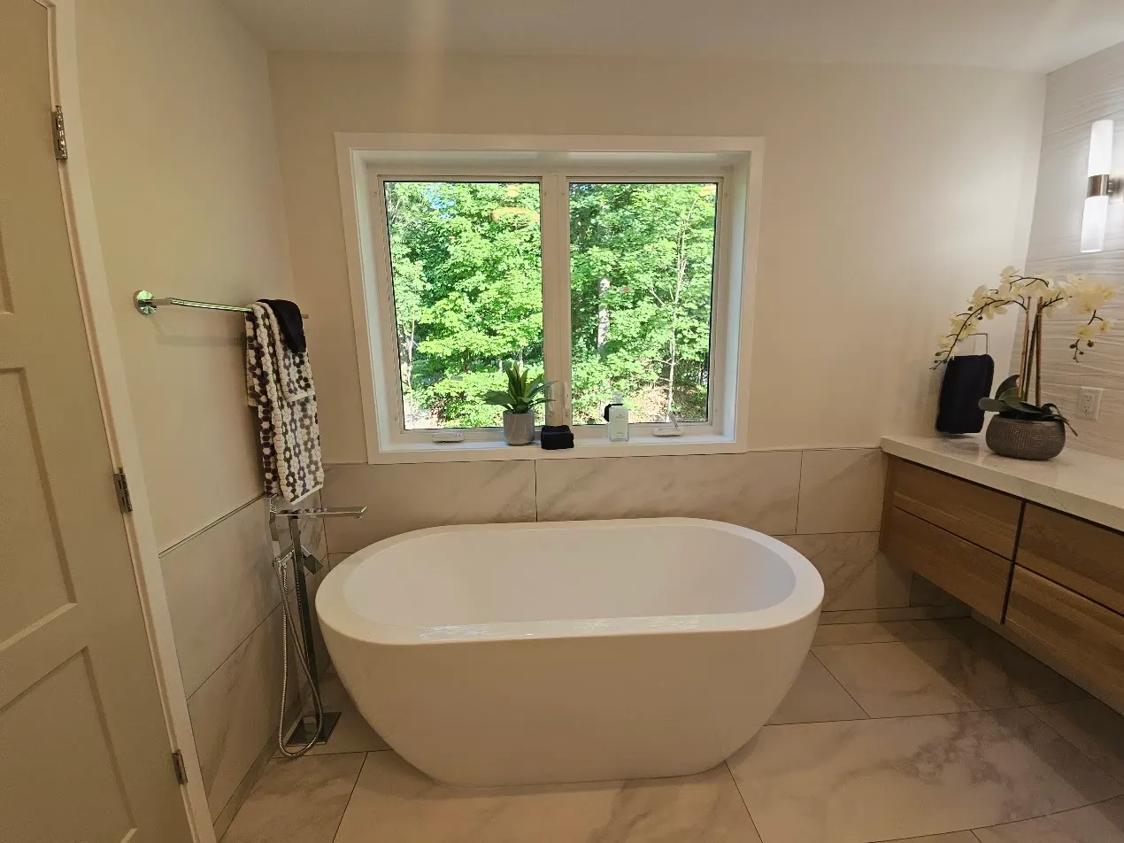 Freestanding white tub with a chrome floor-mount filler under a white-framed window, beside a floating walnut vanity on marble-look porcelain tile. (Filename says "black-window" — the frame is WHITE; name kept because pages already import it. Alt corrected 2026-10-01.)