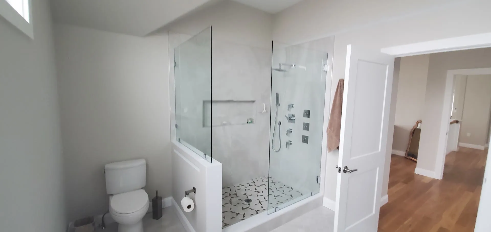 Wide view of a glass shower with multiple body sprays and a pebble floor, a toilet alcove, and a doorway to the hall