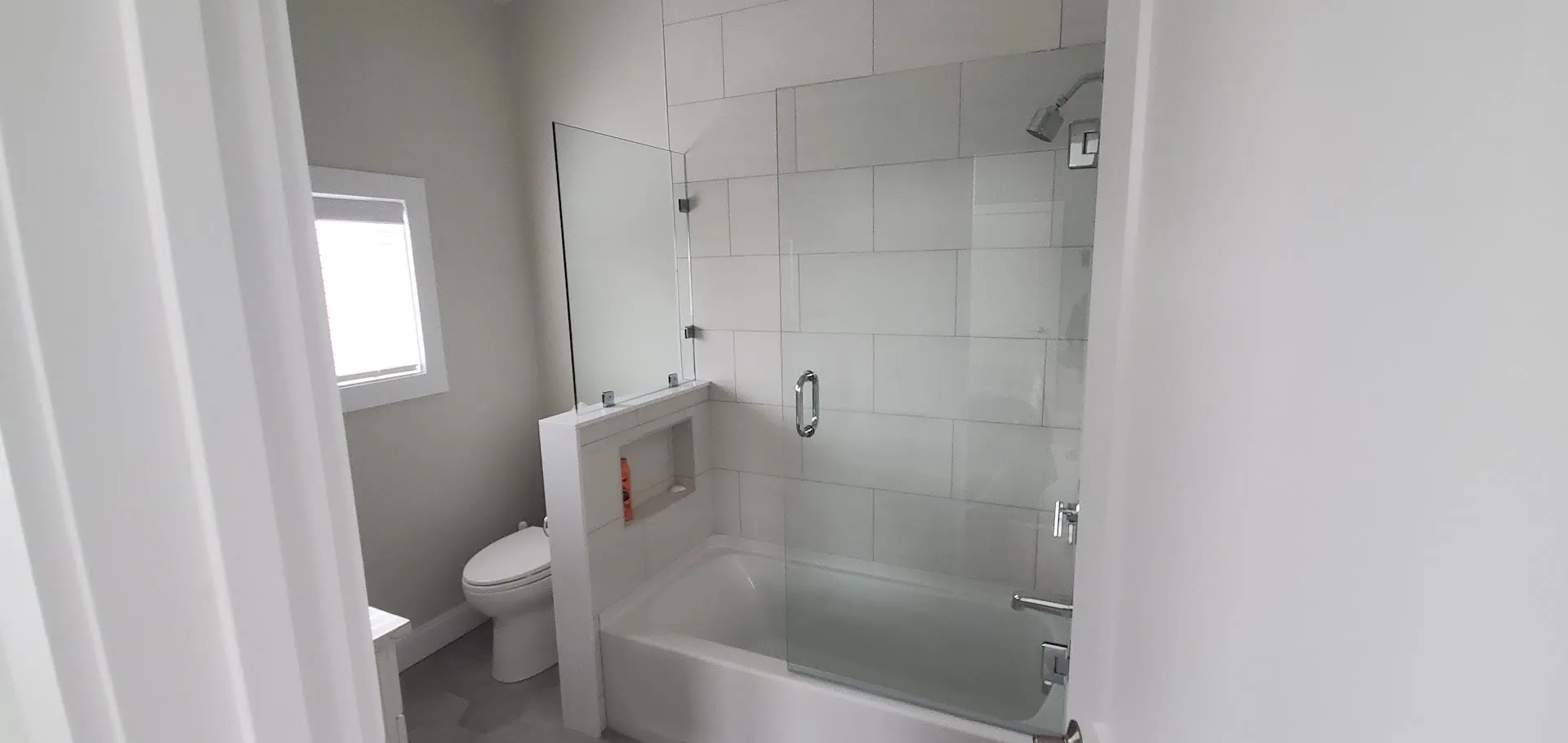 Wide view of a white tub and shower combination with large-format white tile, a glass panel, a tiled niche, and a window
