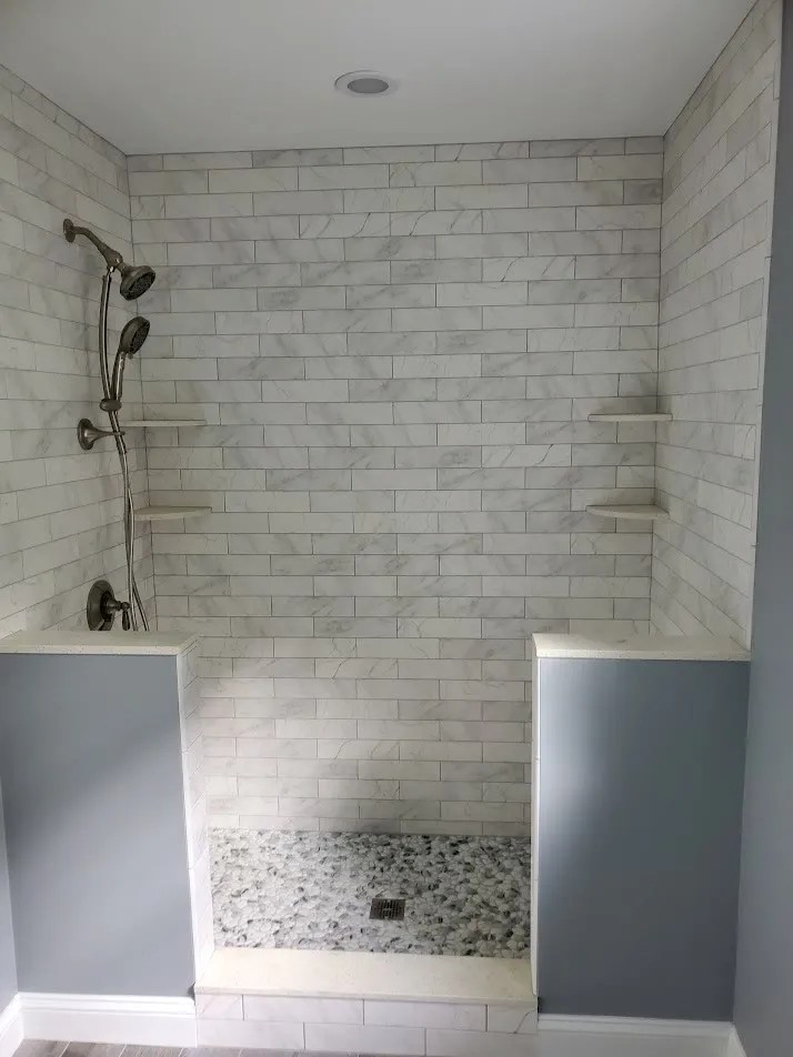 Walk-in shower with marble subway tile, two pony walls, corner shelves, a pebble floor, and oil-rubbed bronze fixtures