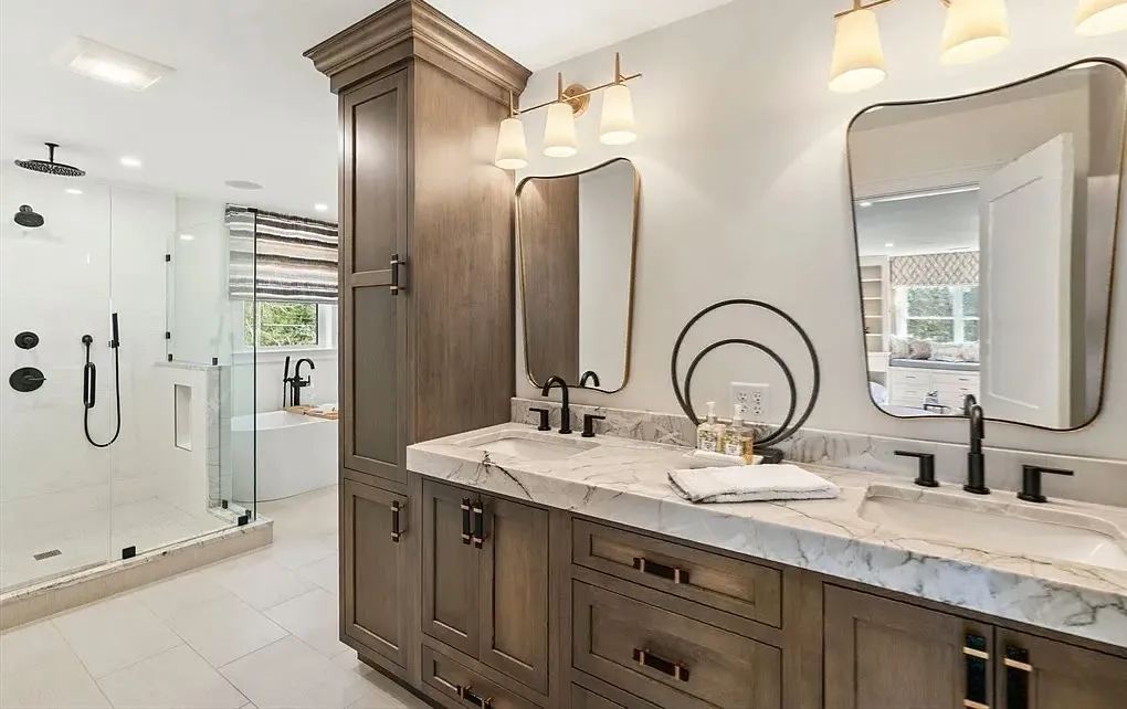 Walnut-stained double vanity and linen tower with a marble-look top, two curved brass-trimmed mirrors, brass sconces, black faucets, and a glass shower with a rain head