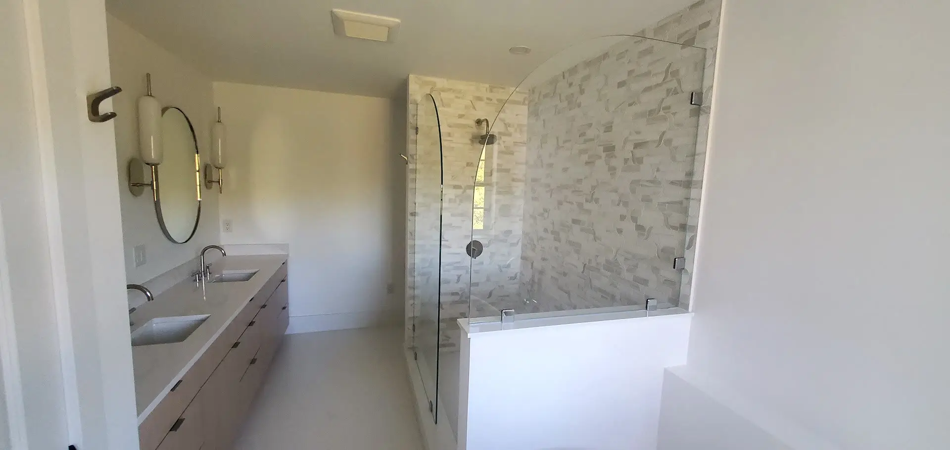 Wide view of a white bathroom with a walnut floating vanity, round mirrors and sconces, and a glass walk-in shower with a stacked-stone accent wall.
