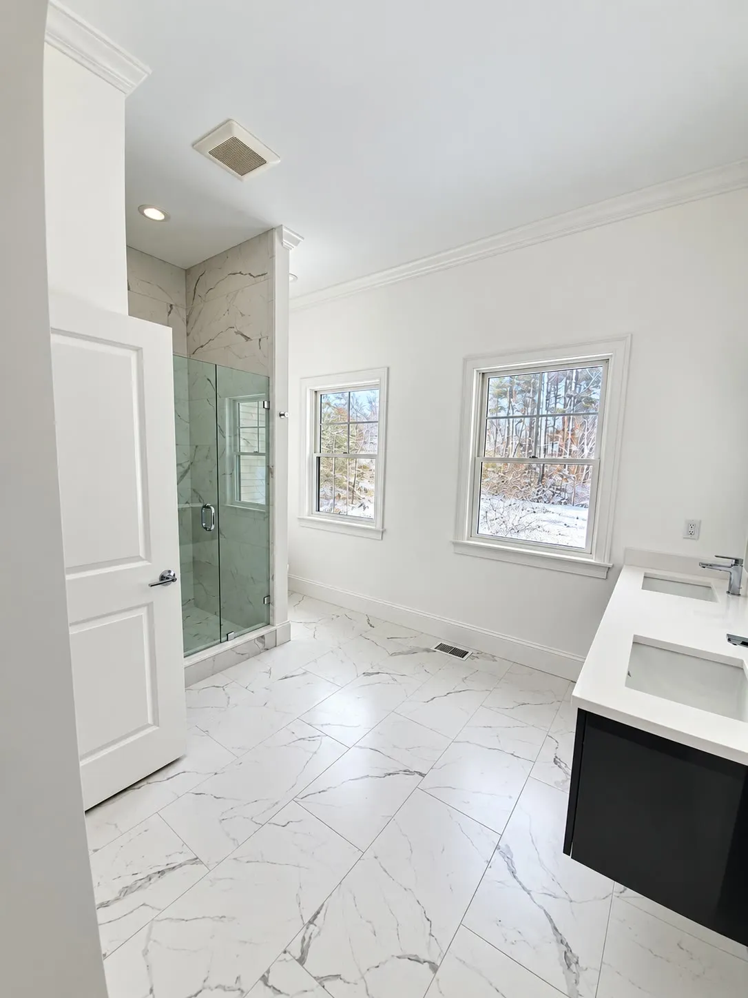 Tall view of a white bathroom with marble-look floors, a glass shower in marble tile, a dark floating vanity with a white top, and two windows