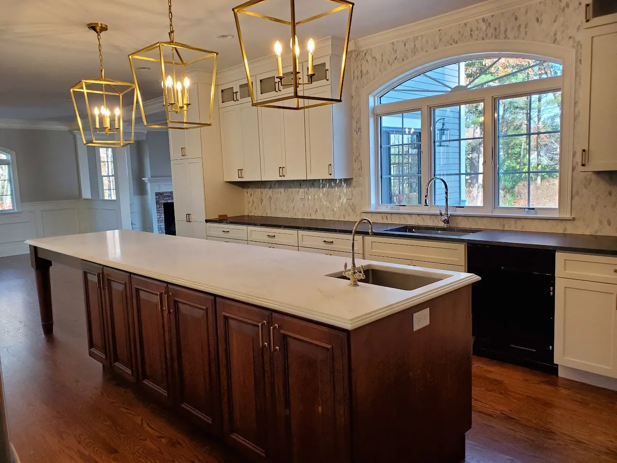 Cherry-stained island with a white quartz top under three brass cage chandeliers, white perimeter cabinets, marble backsplash, and a black-framed arched window