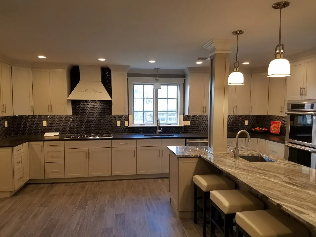 Cream kitchen with a black mosaic tile backsplash, dark granite counters, a raised granite breakfast bar with leather stools, a white custom hood, and double ovens