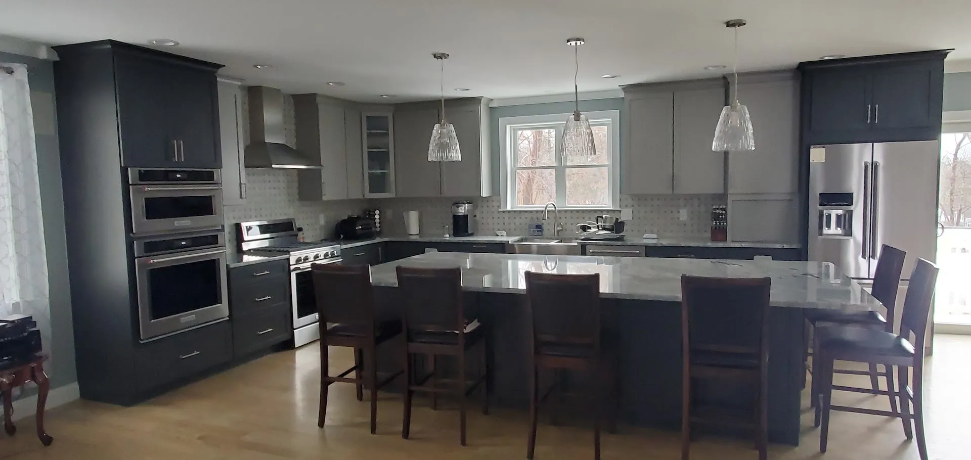 Two-tone kitchen with charcoal tall cabinets, white uppers, a granite island seating six on dark wood stools, stainless hood, and double wall ovens