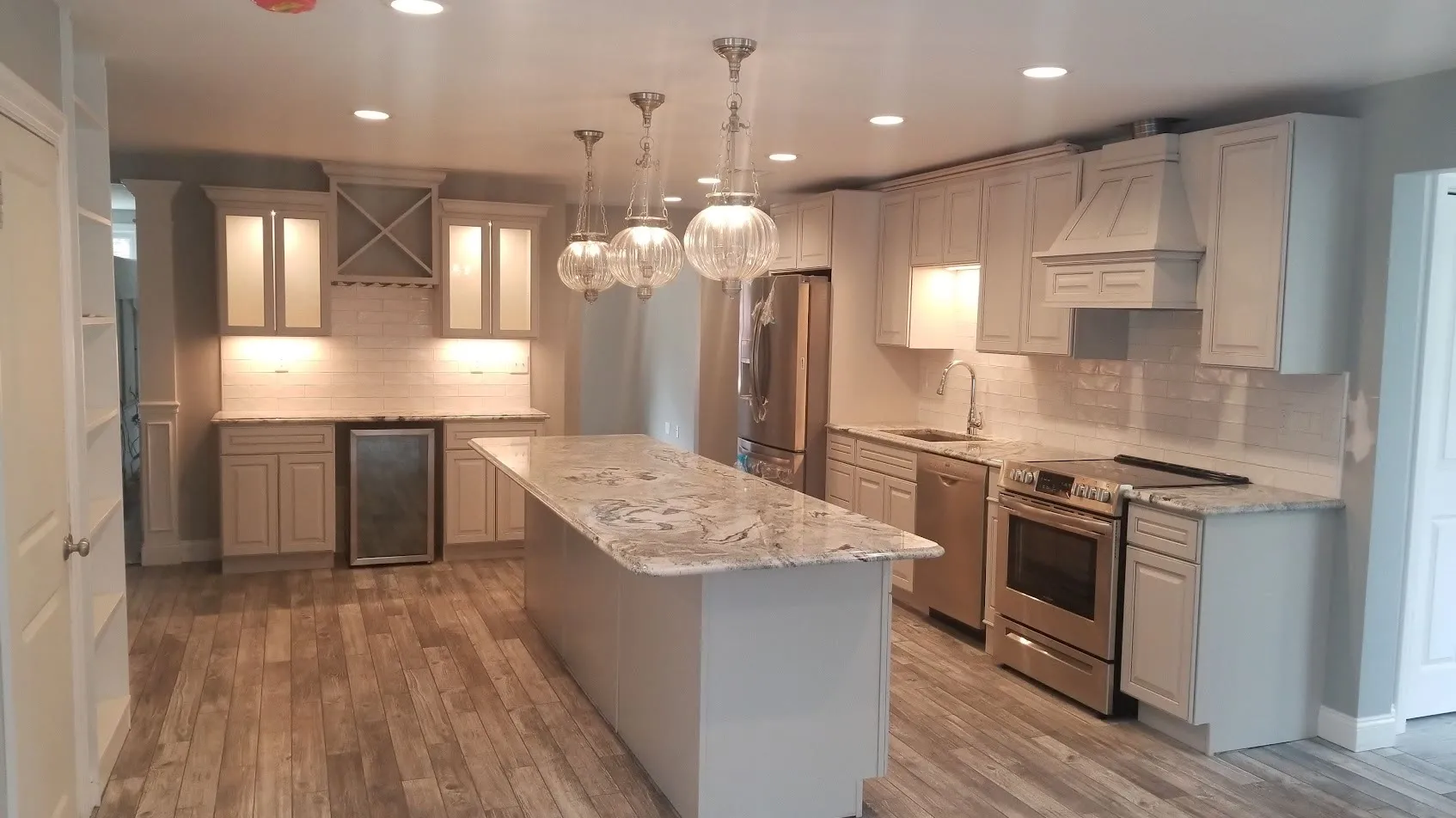 White kitchen with a gray-veined granite island, a cluster of glass globe pendants, white subway tile backsplash, an X-shaped wine cubby, and gray wood-look flooring