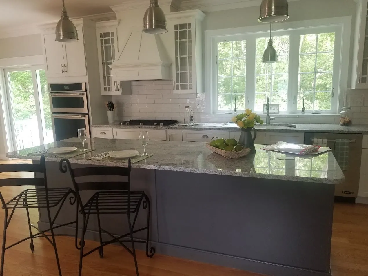 White kitchen with a gray-veined granite peninsula, black wrought-iron stools, white subway tile, a sloped stainless hood, and a large window onto trees