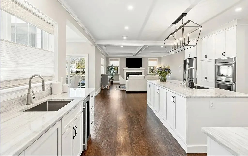 White kitchen with a long marble-look quartz island, a black lantern pendant, coffered ceiling, and dark hardwood floors open to the living room