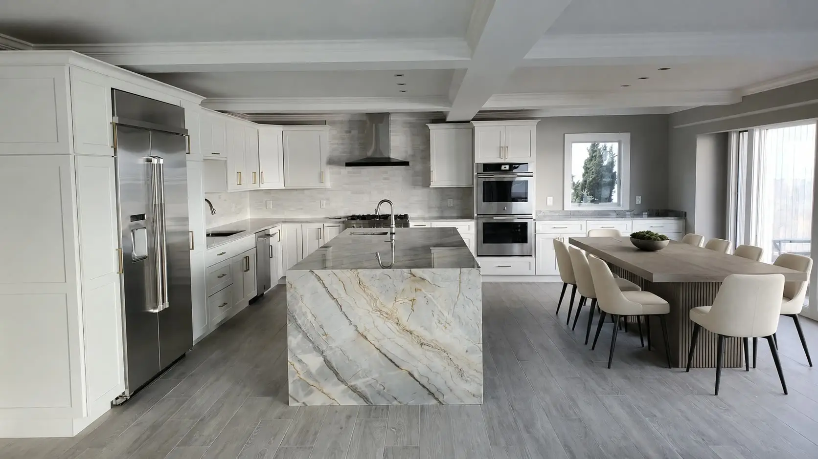 White kitchen with a stainless column refrigerator, a gold-veined quartzite waterfall island, gray wood-look tile floor, and a dining table with cream chairs under a coffered ceiling.