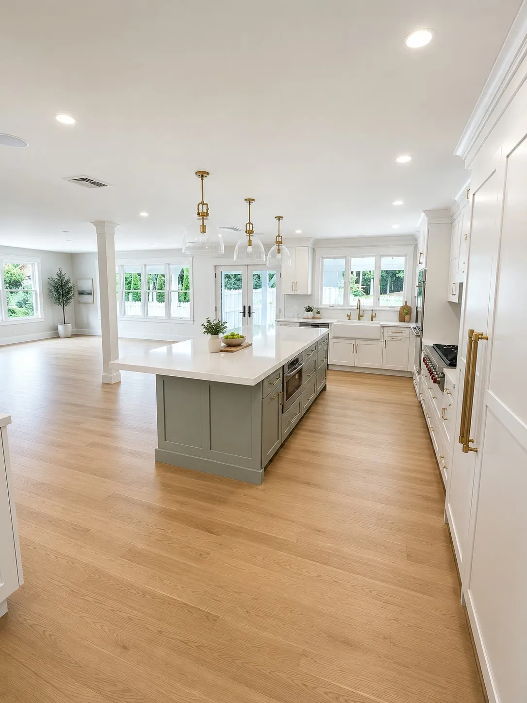 Open-plan white kitchen with a sage-green island, brass hardware, brass and glass pendants, white columns, and wide-plank oak floors running into the living area