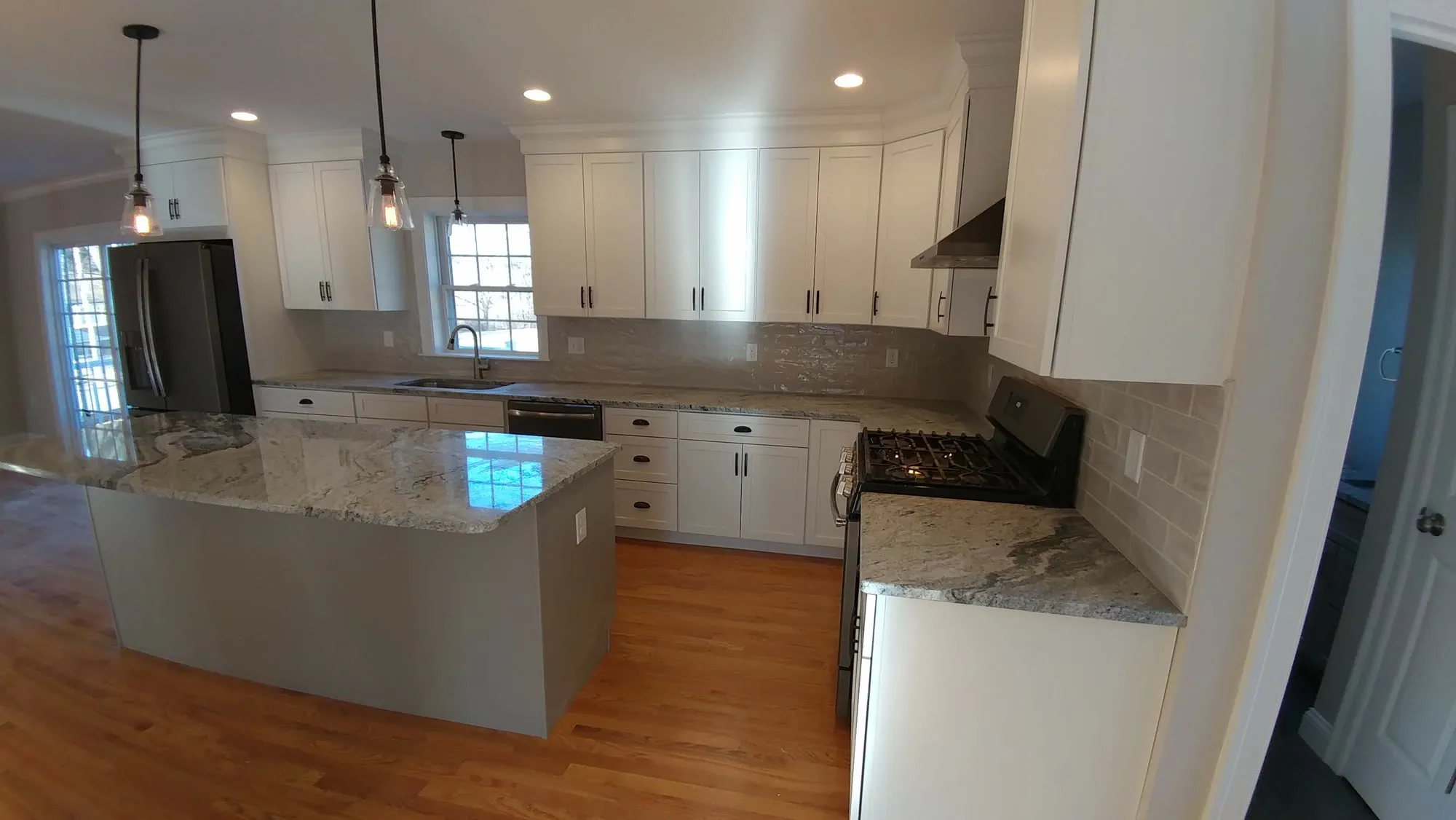 Large white shaker kitchen with a gray-based granite island, gray subway backsplash, stainless range, two windows, and three glass pendants over hardwood floors