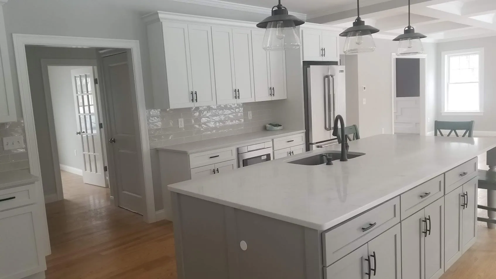 View through a doorway into a white shaker kitchen with a gray island, white quartz counters, black pendants, and a black faucet on hardwood floors
