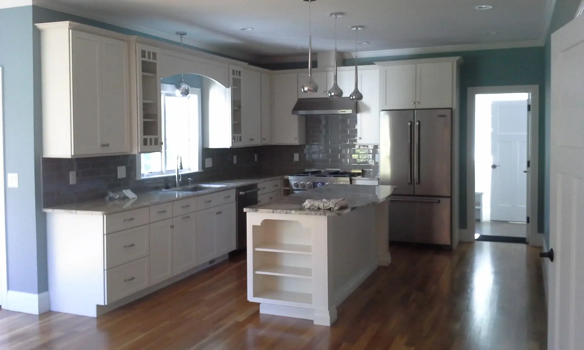 White shaker kitchen with gray subway tile backsplash, stainless range and hood, and a granite-topped island with open end shelving on hardwood floors.