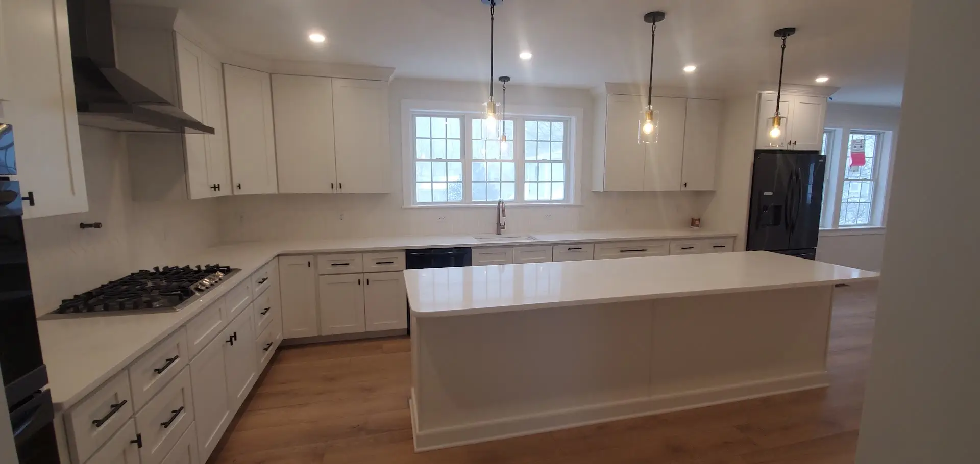 White shaker kitchen with an oversized white quartz island, a gas cooktop, stainless range hood, and three filament pendants over hardwood floors.