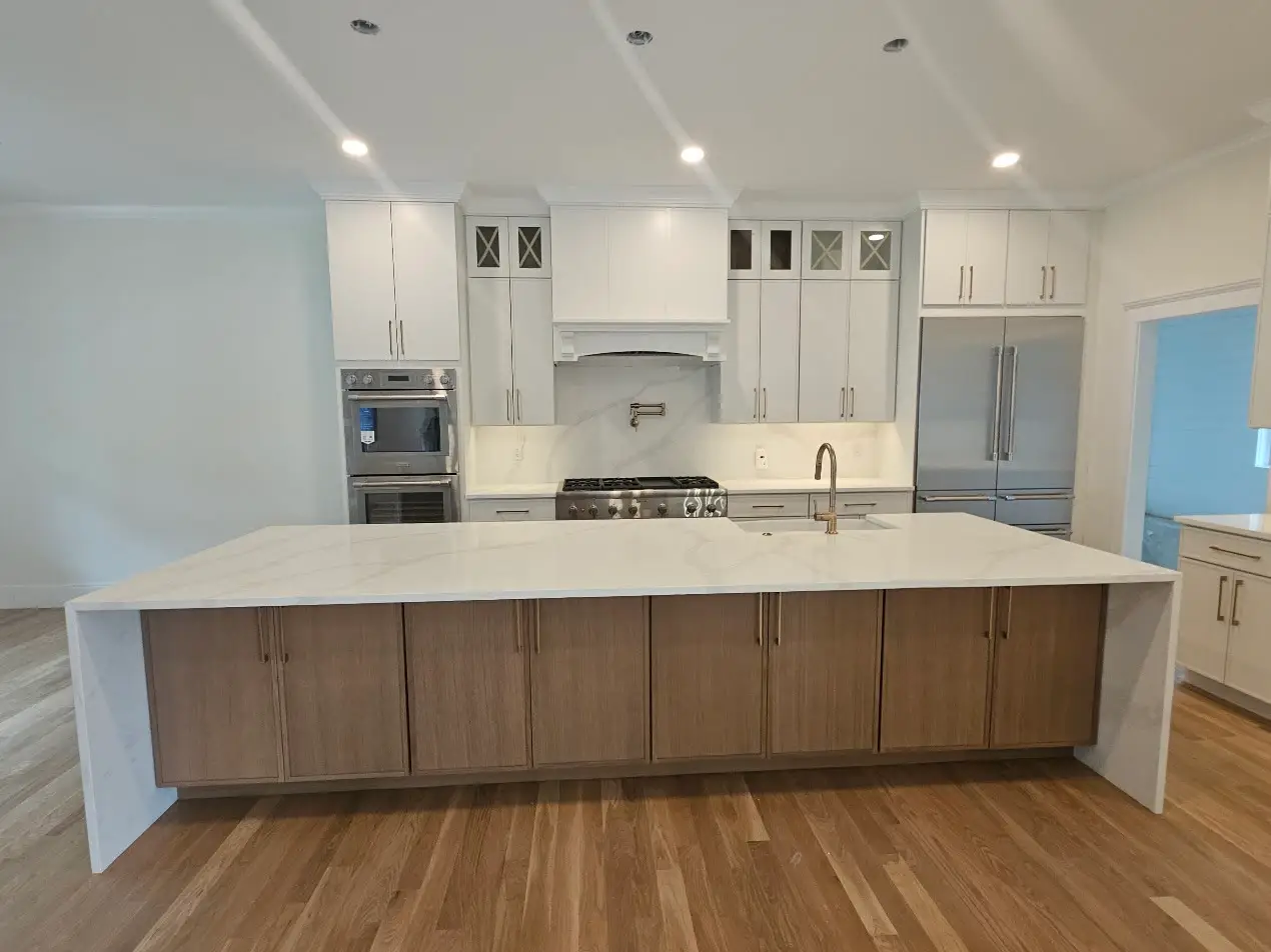 Long oak-base island with a white marble-look quartz waterfall top, white slab cabinets, a pot filler over the range, stainless double ovens, and wide-plank oak floors.