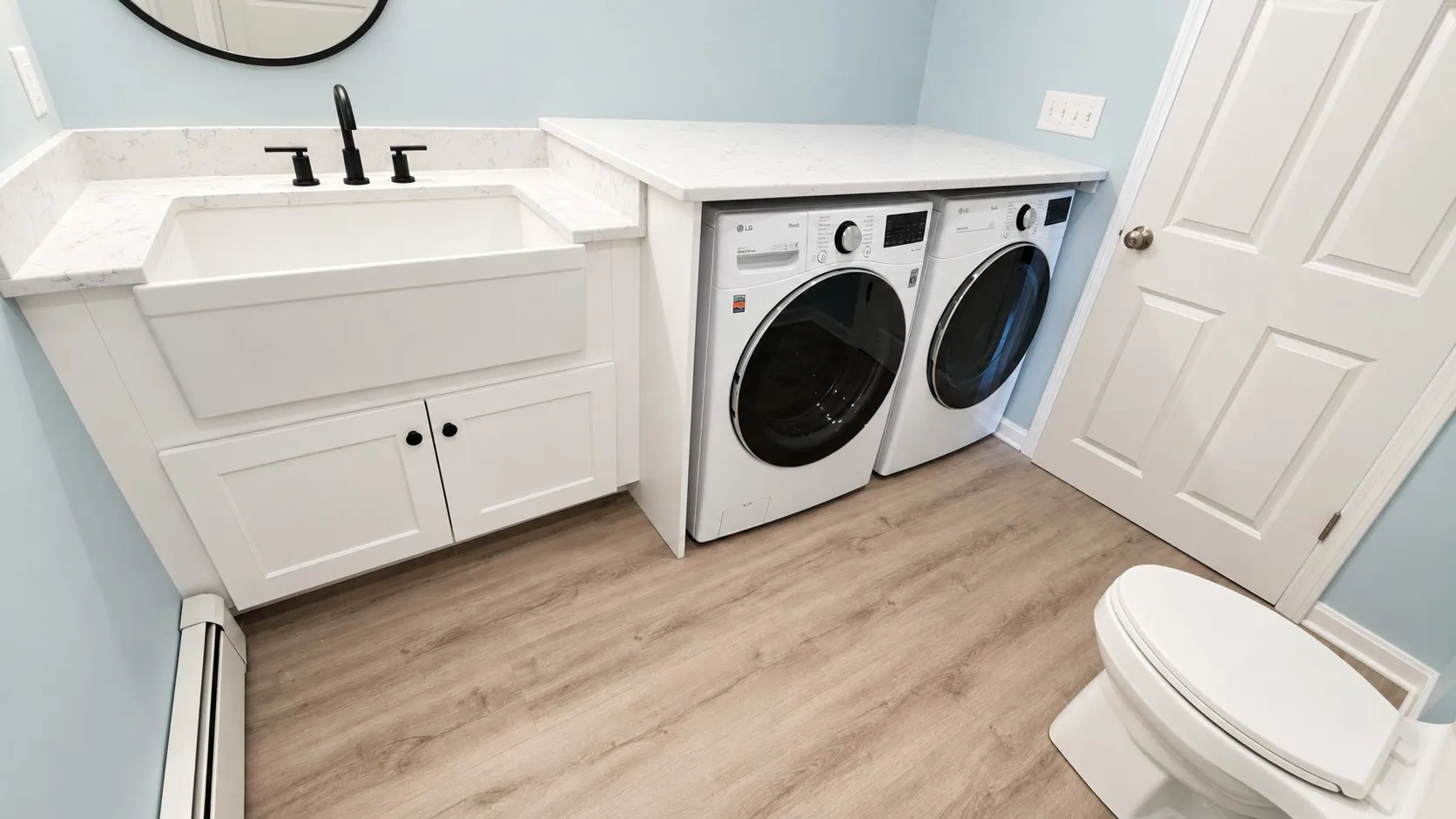 Laundry room with a white farmhouse utility sink and cabinet, marble-look counter, black faucet, front-load washer and dryer, light blue walls, and oak-look floors