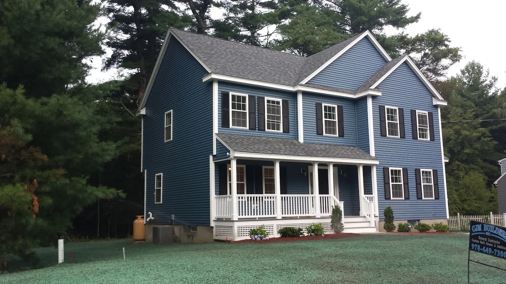 Blue colonial with a white farmer’s porch and twin front gables on a freshly hydroseeded lawn, with a CJM Builders yard sign