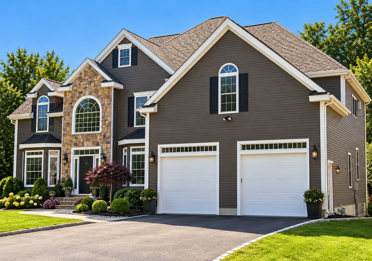 Brown two-story home with stone veneer accents, arched windows, a two-car garage with white doors, and a Japanese maple