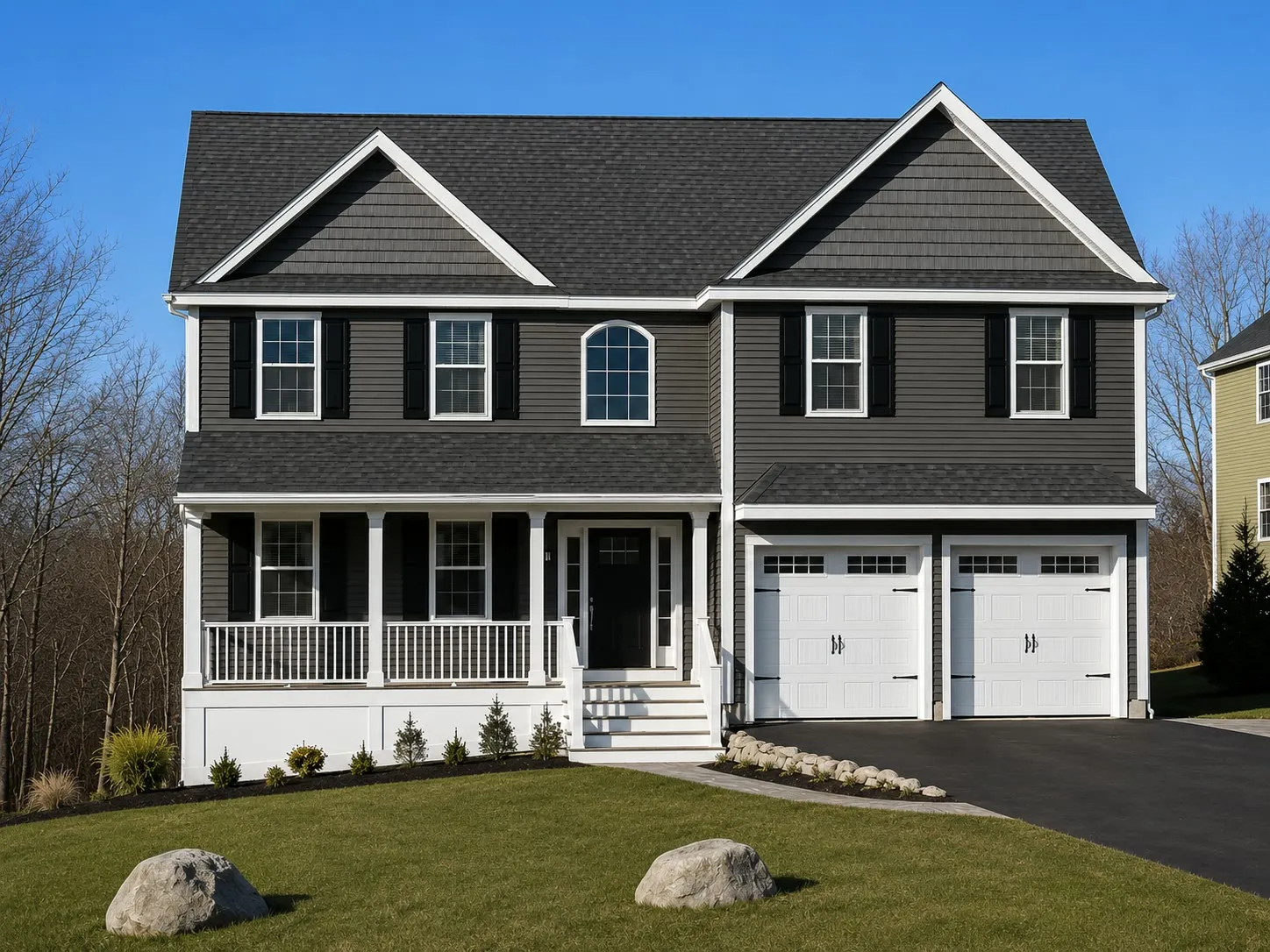 Charcoal two-story colonial with shake gables, a white farmer’s porch, a two-car garage with white doors, and boulders in a new lawn