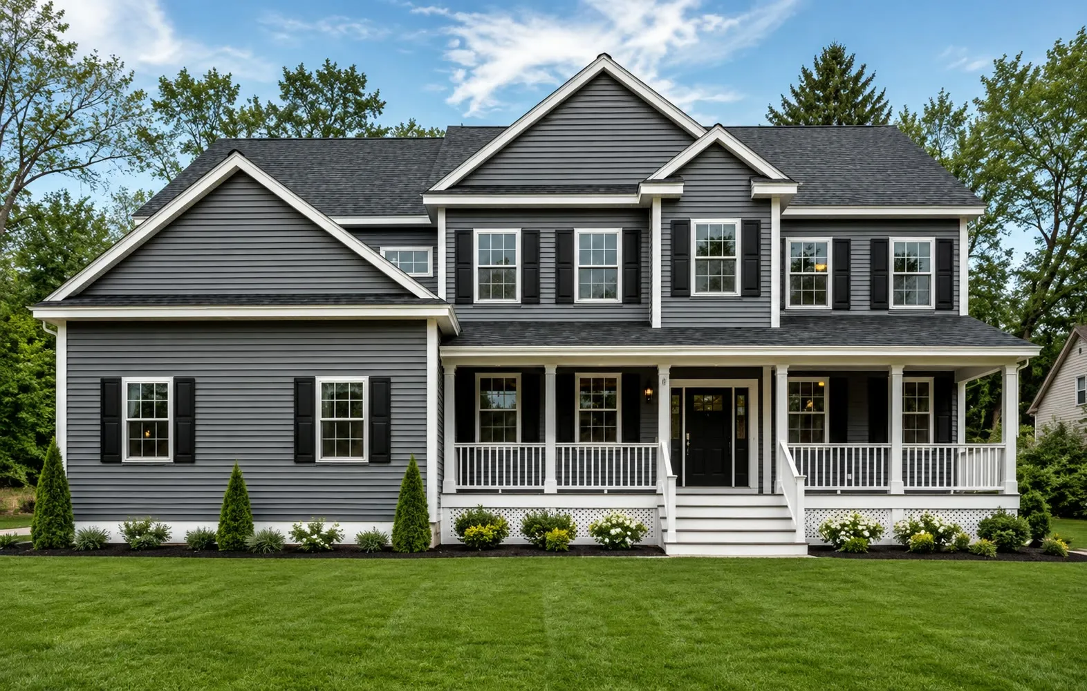 Gray colonial with black shutters, a white farmer’s porch, and a second front gable, with evergreens and a manicured lawn