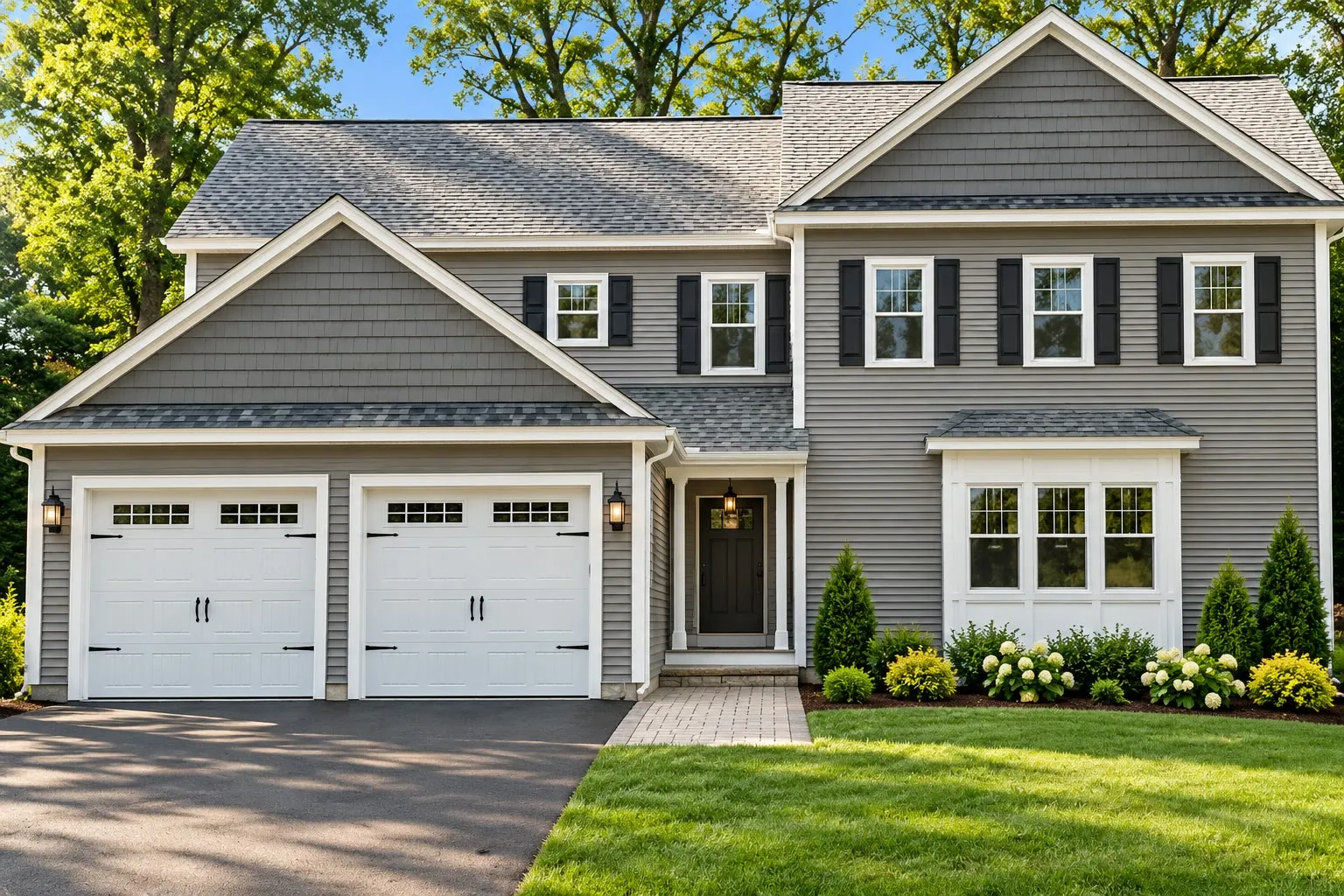 Gray colonial with a cedar-shake gable, black shutters, a bay window, a two-car garage with white carriage doors, and hydrangeas along a paver walk