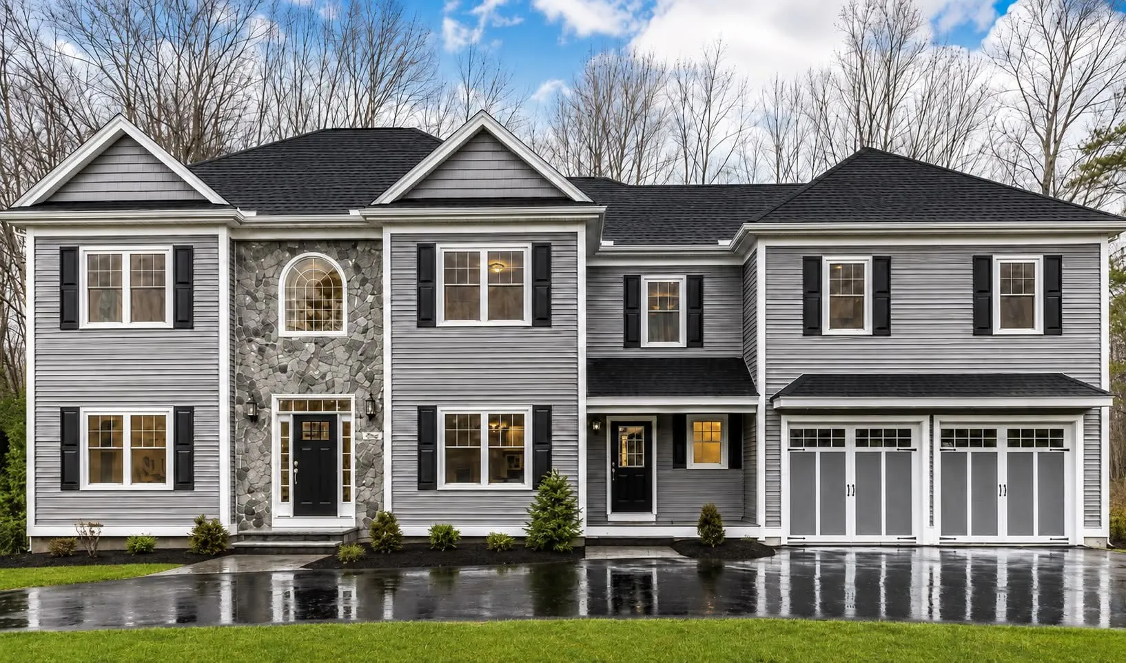 Gray colonial with a stone-veneer entry, an arched window, black shutters, and a two-car garage with carriage doors on a wet driveway