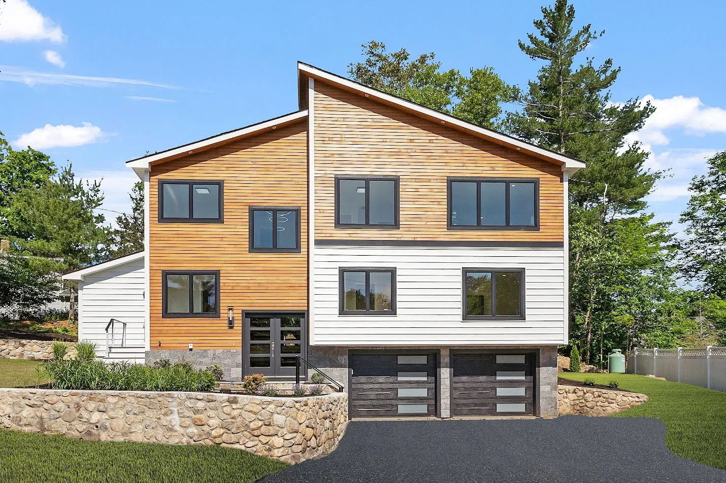 Contemporary shed-roof home with cedar siding above white, black windows, a stone retaining wall, and a drive-under garage
