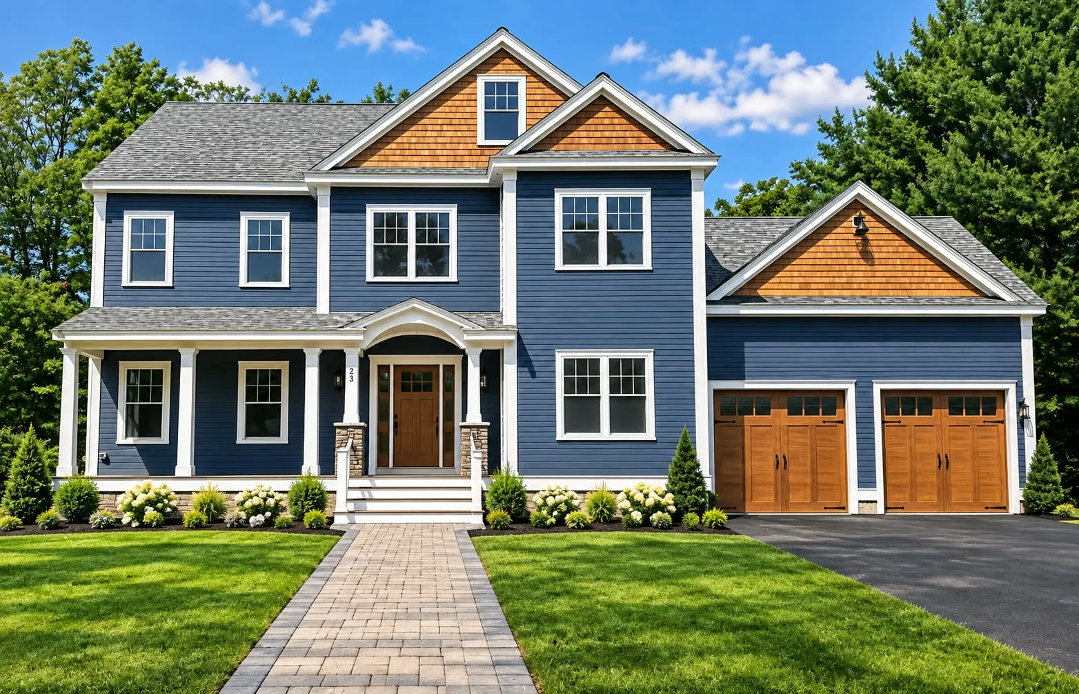 Navy colonial with cedar-shake gables, a white farmer’s porch on stone piers, wood carriage-style garage doors, and a paver walkway