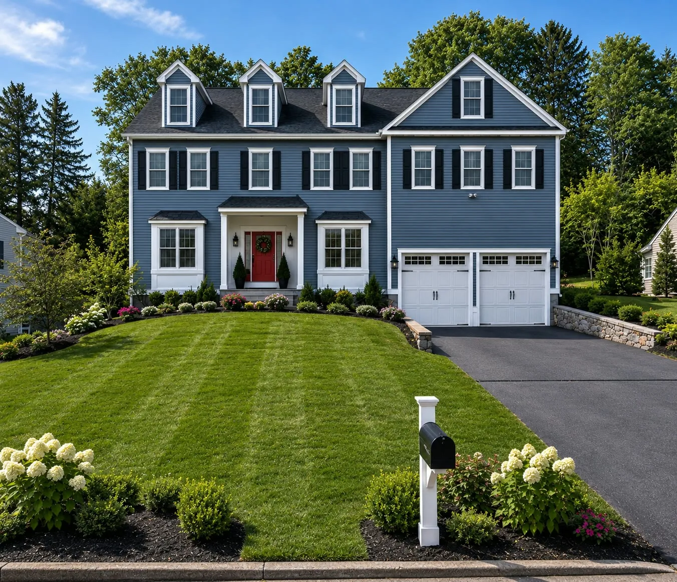 Slate-blue colonial with three dormers, black shutters, a red front door, a two-car white garage, and hydrangeas by the mailbox