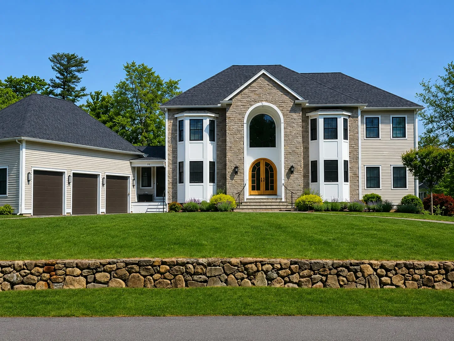Tan two-story home with a two-story arched entry, stone-veneer accents, a three-car garage, and a fieldstone wall