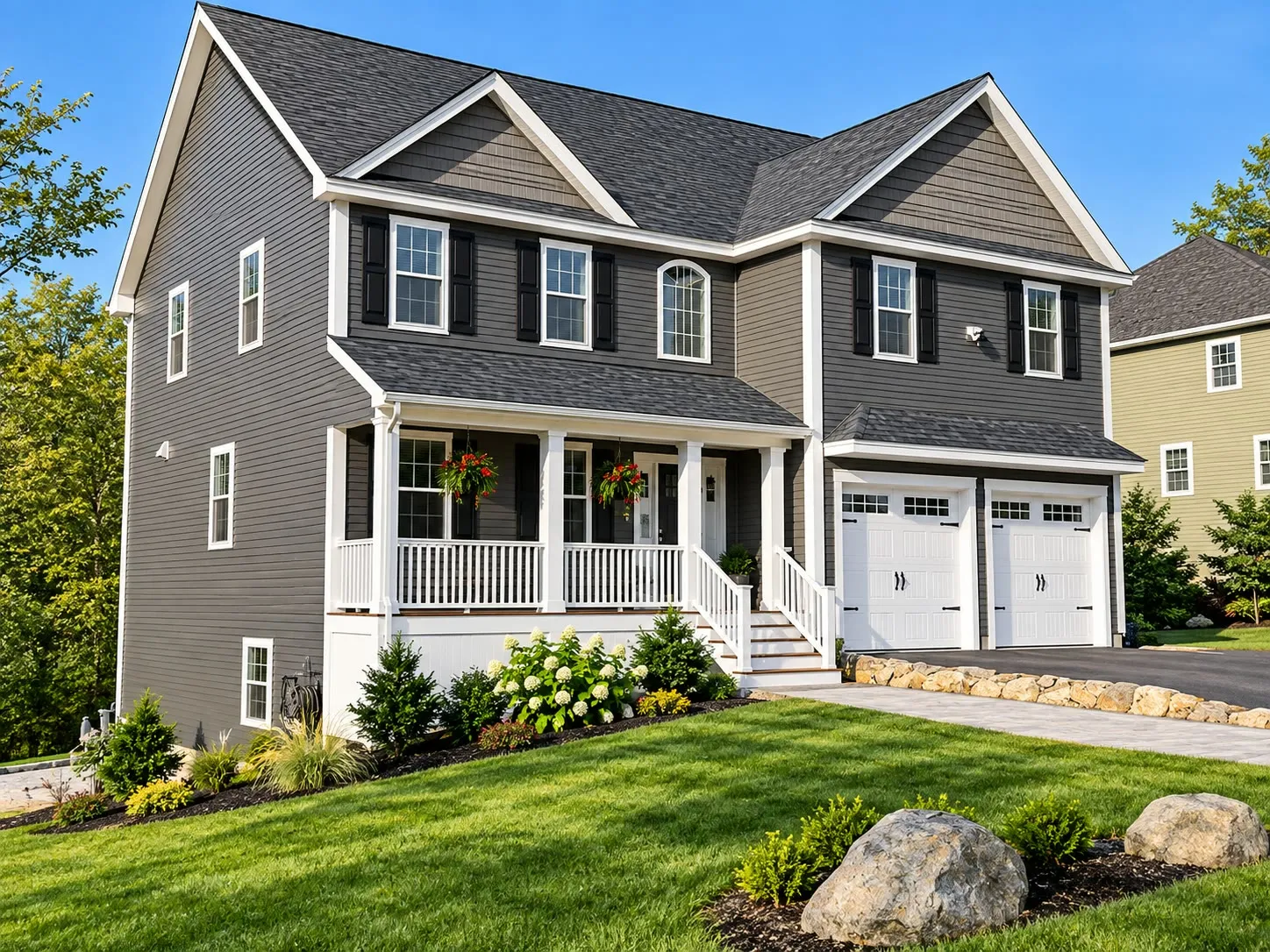 Taupe two-story home with shake gables, a farmer’s porch, a two-car garage, a stone wall, and boulders in the landscaping