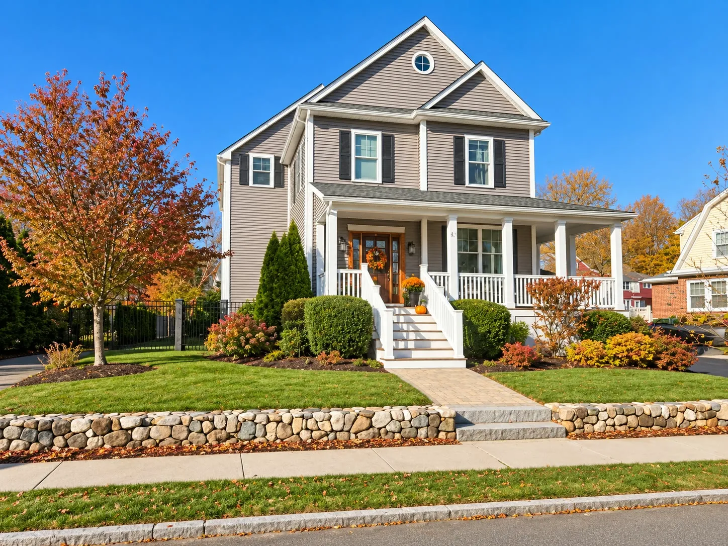 Taupe two-story home with a farmer’s porch and a round attic window behind a stone wall, with autumn foliage