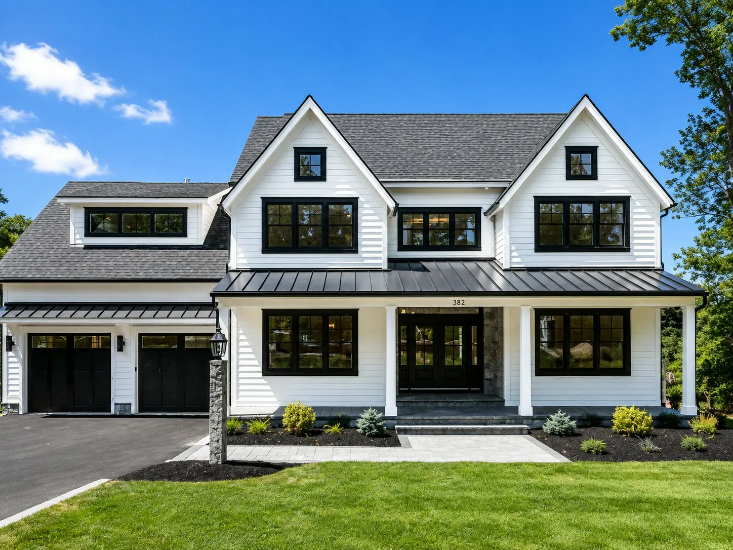 White modern farmhouse with black windows, a black standing-seam porch roof, a stone column base, black garage doors, and house number 382 on the porch