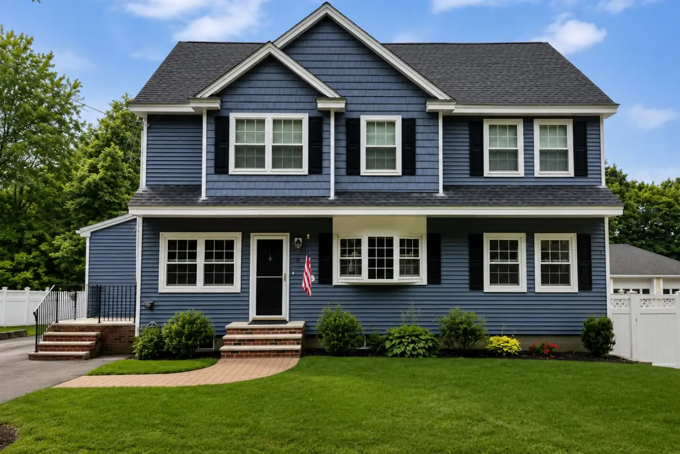 Blue two-story colonial with shake gables, black shutters, a brick walkway, and a white fence on the same lot in summer
