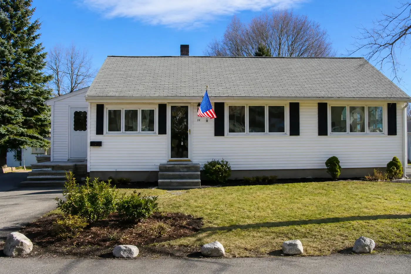White single-story ranch with black shutters, an American flag, and boulders along the lawn in early spring