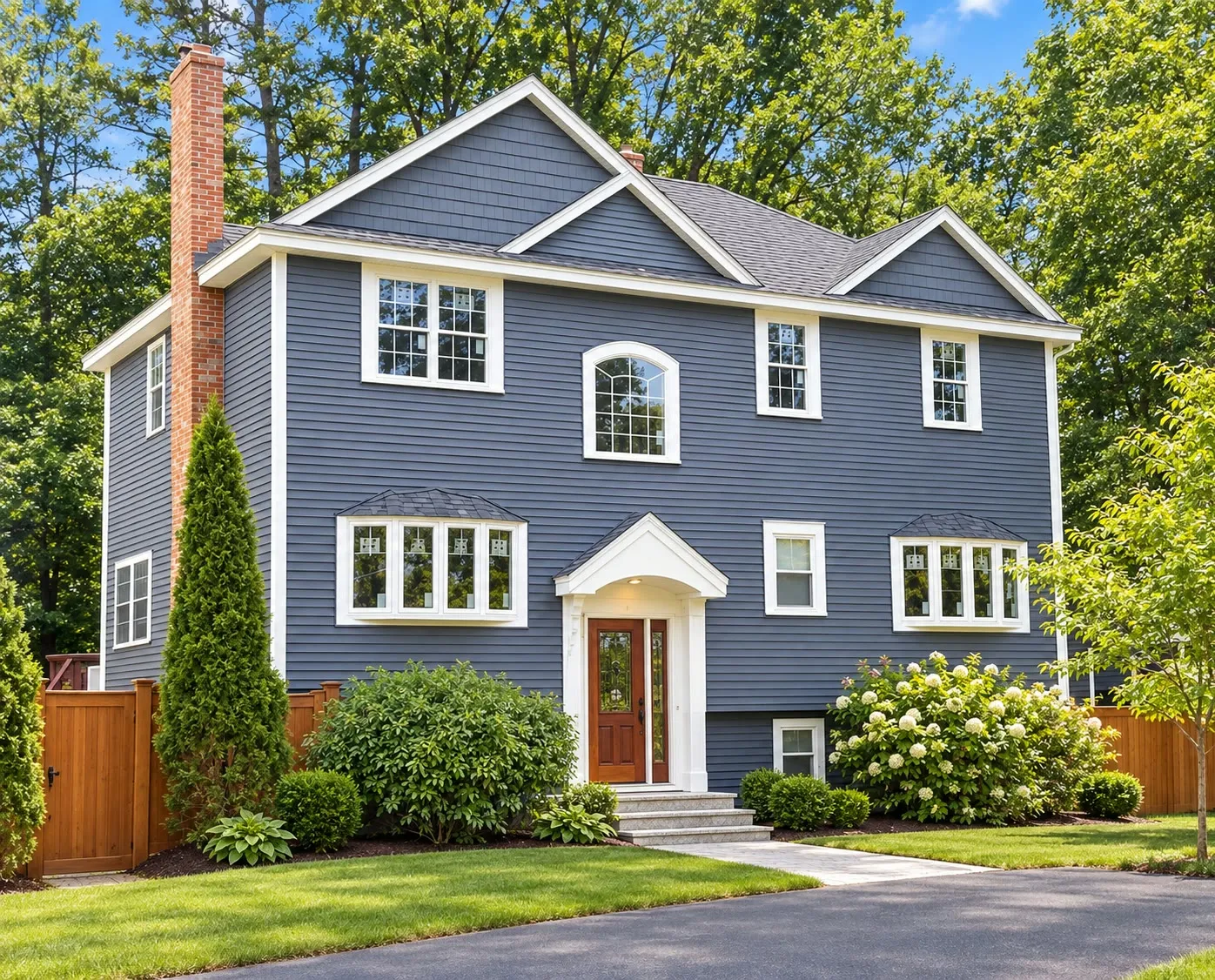 Blue-gray two-story home with a brick chimney, an arched window over the entry, a red front door, white trim, hydrangeas, and a cedar fence