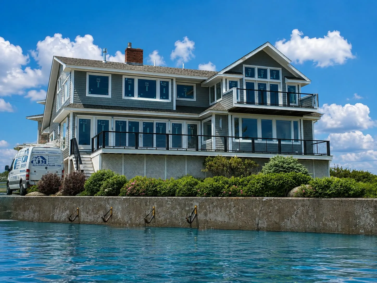 Gray cedar-shingle waterfront home with two levels of decks and black railings above a stone seawall, with a company van parked at left