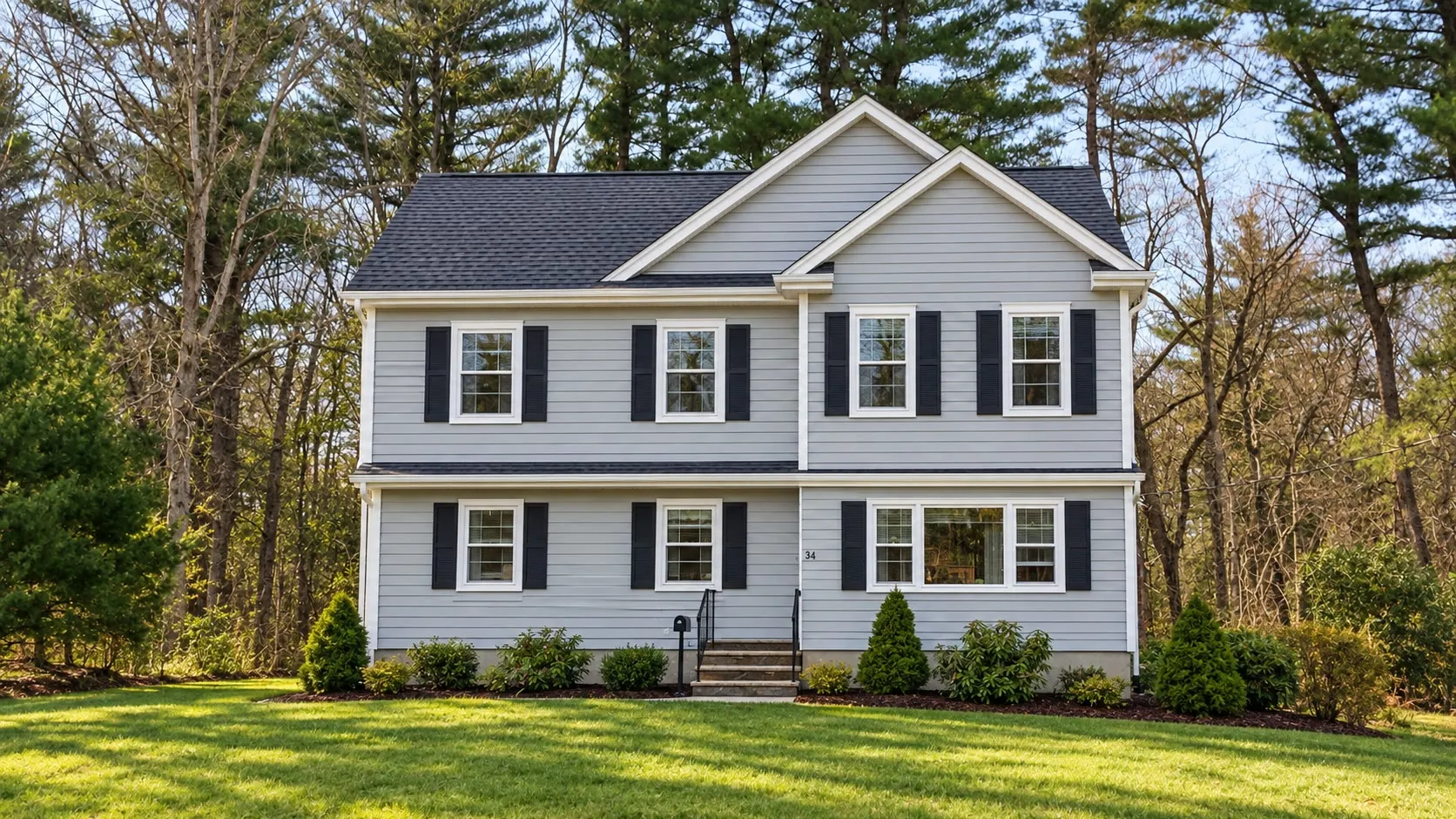 Light gray two-story colonial with black shutters, a front gable, white trim, and a fresh lawn against a wooded backdrop