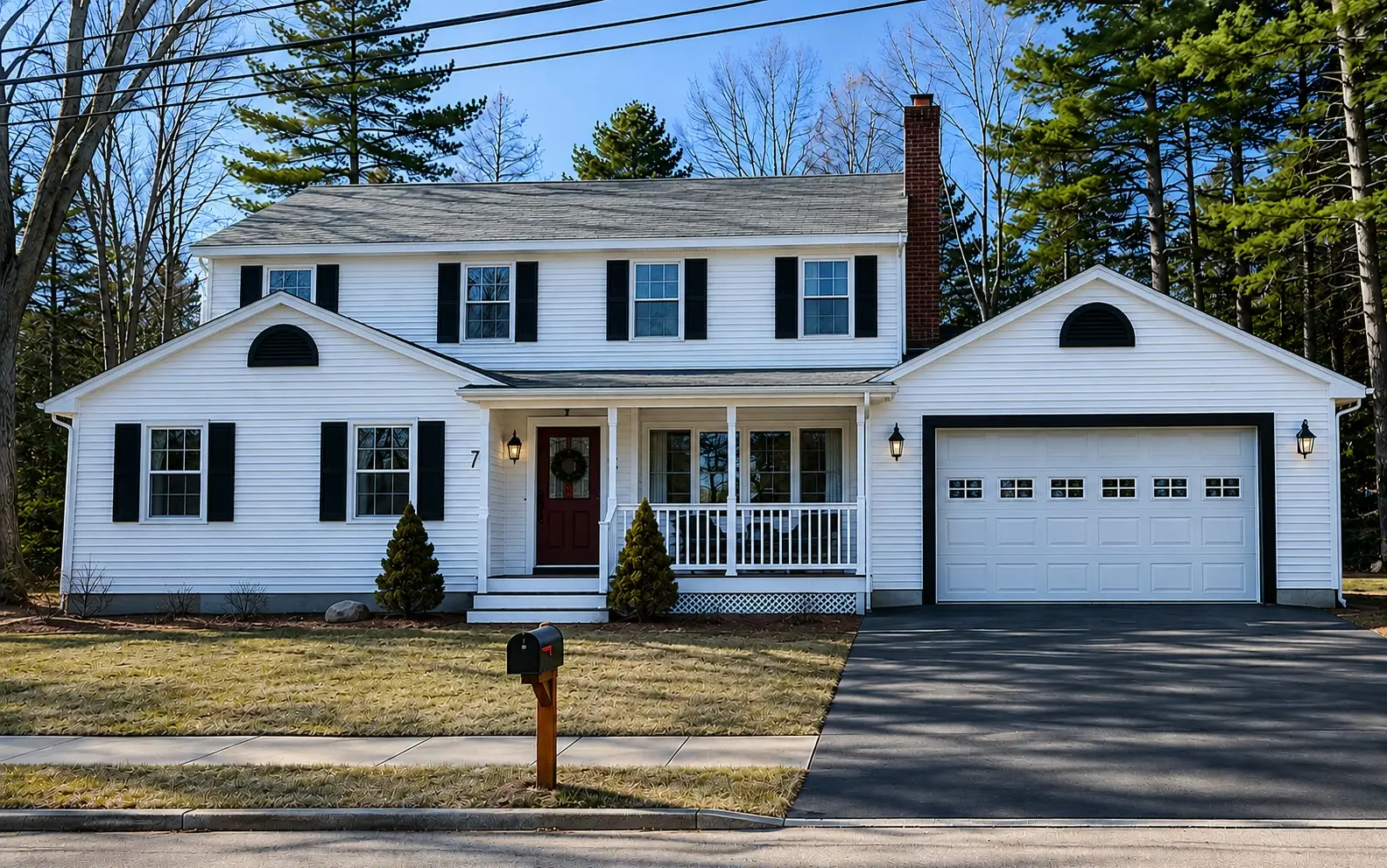 White colonial with black shutters, a red front door with a wreath, a farmer’s porch, a brick chimney, and an attached garage with a half-round window.